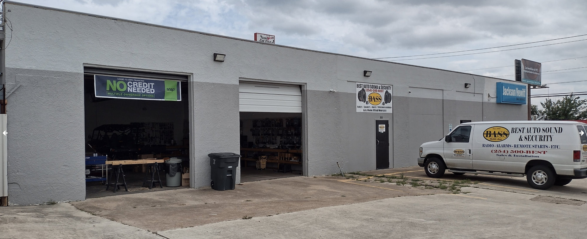 A white commercial service van with Best Auto Sound & Security branding parked in front of a gray industrial building with open garage doors.