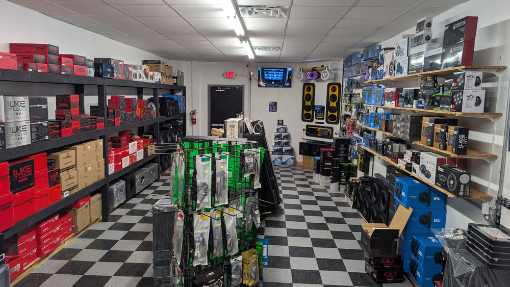 A retail electronics store interior with shelves and racks filled with boxed electronic hardware, speakers, and accessories. The floor is black-and-white checkered, and the store is lit by overhead LED lights.