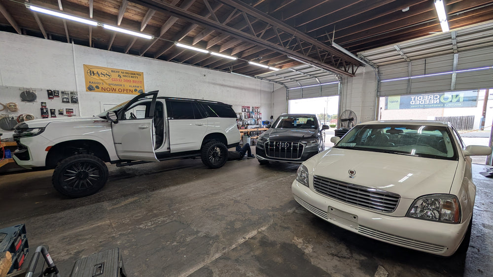 A white Cadillac sedan parked inside a garage next to a white lifted SUV with its door open, and a dark Audi SUV in the background.