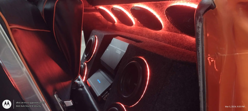 Custom car interior sound system with a custom orange door panel, black leather seats, and a custom carpeted footwell illuminated by red LED accent lighting.