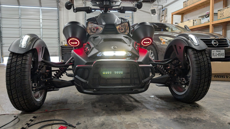 A black Can-Am three-wheeler vehicle photographed from the front in a garage setting, with its headlights and red DS18 circular Speaker pod rings illuminated. A gray Nissan SUV is parked in the background.