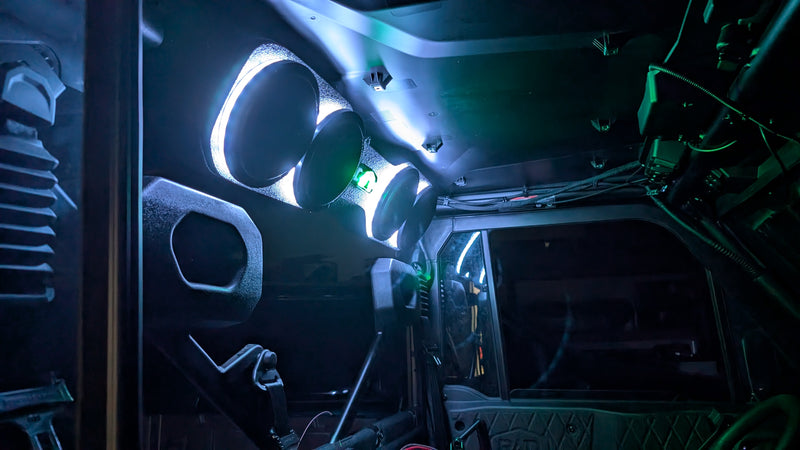 A dark vehicle cabin interior illuminated by glowing green and blue LED light strips, showing the dashboard, steering wheel, and cabin ceiling.