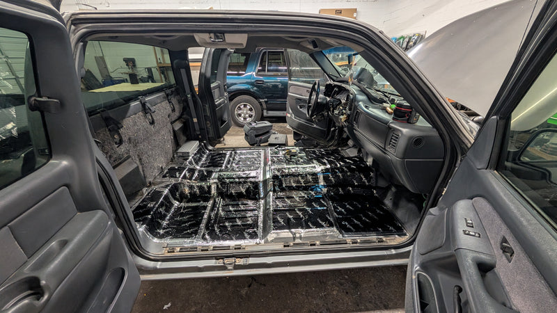 The open rear doors of a dark gray Chevy truck showing a stripped-out interior with the carpet panel removed, exposing a black sound deadening material. The vehicle is parked inside a workshop or garage with another blue vehicle visible in the background.
