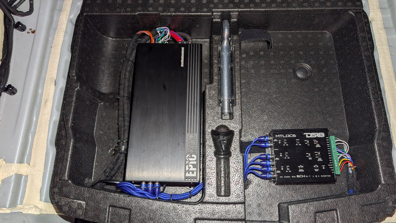 An automotive audio upgrade kit displayed inside a custom black foam tray, featuring a large black amplifier, a control box with blue wiring, a speaker wire, and tools.