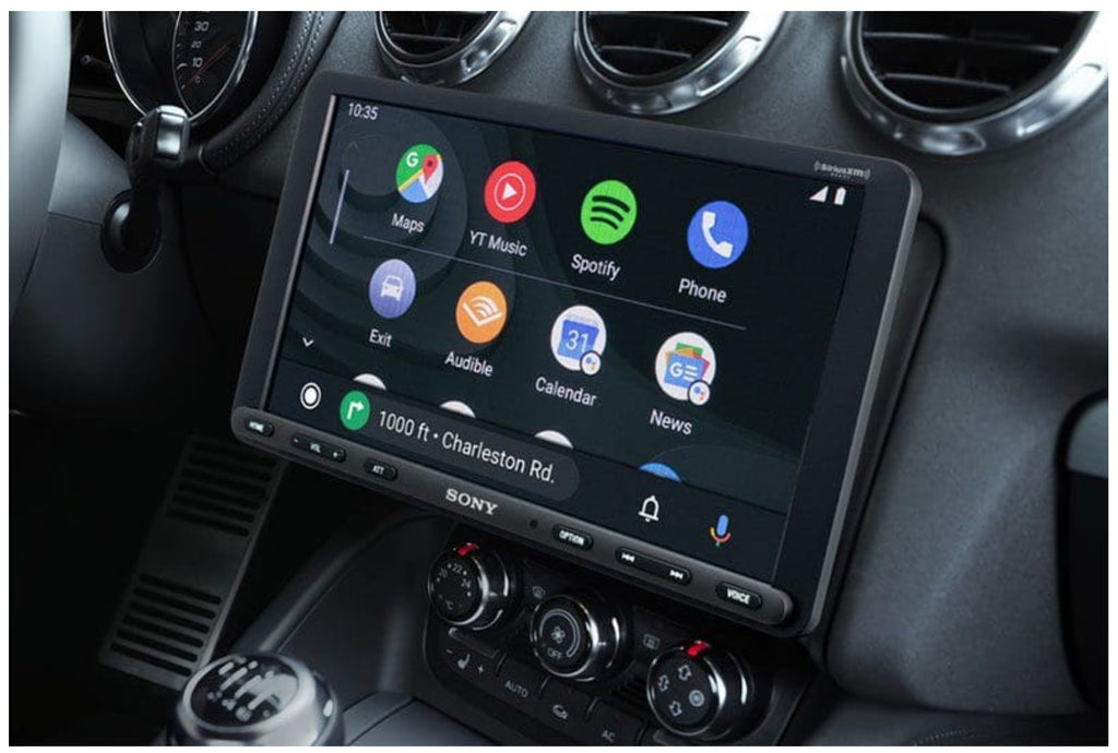 Sony XAV-AX8100 installed in vehicle dash showing Android Auto interface with Maps, YouTube Music, Spotify, Phone, Audible, Calendar, and News apps with turn-by-turn navigation to Charleston Rd