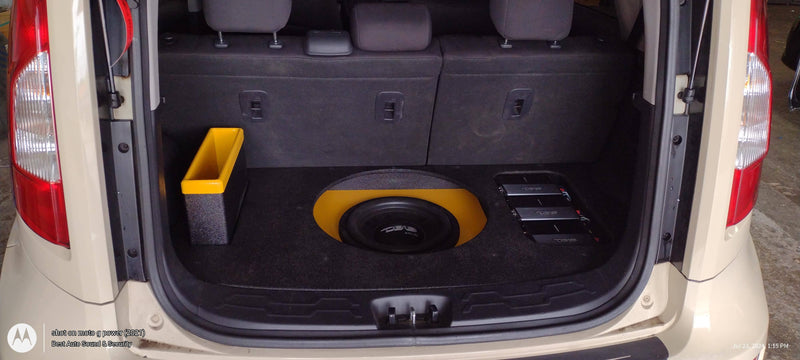 Open trunk of a beige Kia Soul showing a large circular subwoofer installed in the cargo area, with a yellow accent around the speaker and a yellow storage bin on the left.