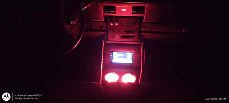 A car interior console illuminated by red ambient lighting, showing a center console with a small screen and two glowing cup holders 